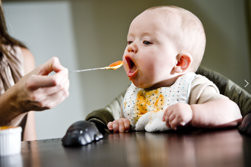 Baby in highchair being fed an orange puree on a spoon