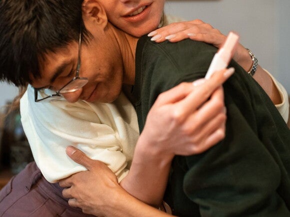 man in green shirt and woman in white shirt hugging with a pregnancy stick in hand