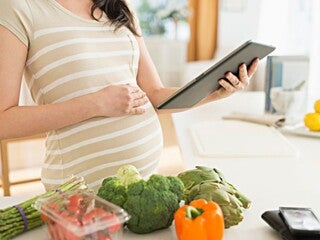 Pregnant woman preparing food and reading from an iPad.