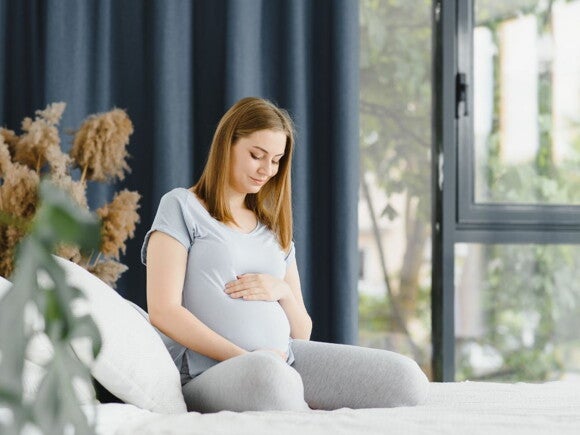 Pregnant woman in grey top and pants sitting on her bed holding her belly