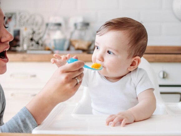 Baby wearing white in highchair being fed puree by a parent on a spoon