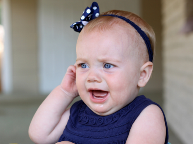 Young infant in blue dress crying and pulling on ear
